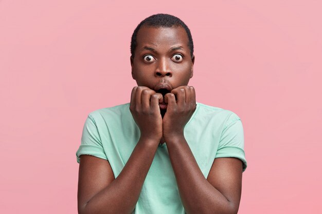 Surprised nervous dark skinned male student bites finger nails, stares at camera with bugged eyes, being afraid of difficulties to pass exam, dressed casually