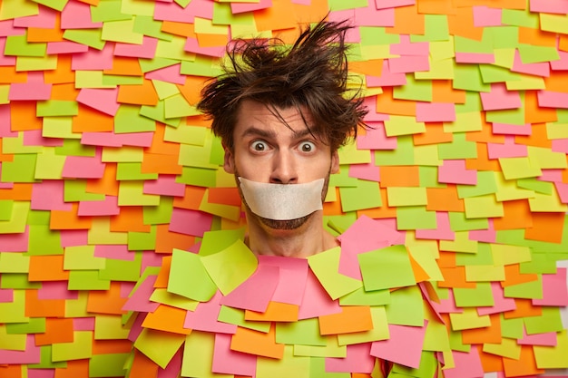 Surprised nervous man with adhesive duct tape over mouth, asks be silent, stands silenced and speechless, poses against colorful wall with sticky notes, being frightened. Shut up, censorship