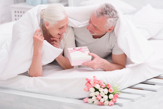 Surprised old woman looking at gift box given by her husband on bed