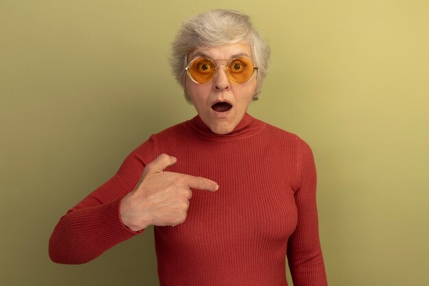 Surprised old woman wearing red turtleneck sweater and sunglasses pointing at space in front of her isolated on olive green wall with copy space