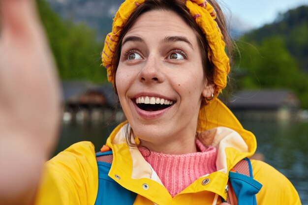 Surprised optimistic young woman has toothy smile, makes selfie, focused somewhere aside