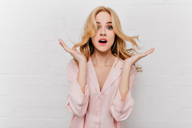 Surprised pale girl posing on white wall. Indoor photo of shocked caucasian woman in cute silk night-suit.