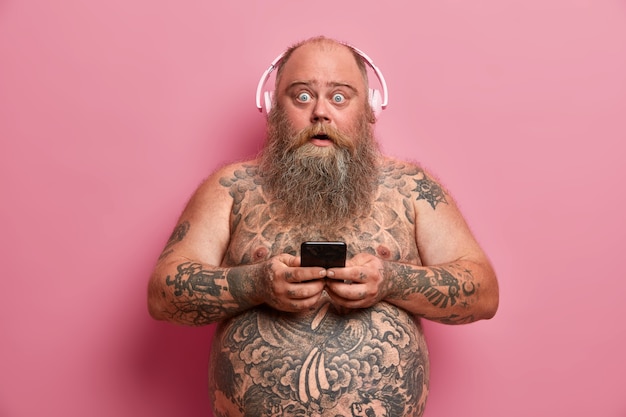 Surprised plump man stares and says wow, texts with friend via smartphone, wears headphones on ears, listens music, poses shirtless, has tattooed body. Technology and leisure concept