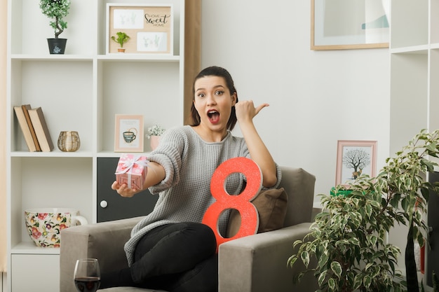 Surprised points at side beautiful girl on happy women day holding out present at camera sitting on armchair in living room