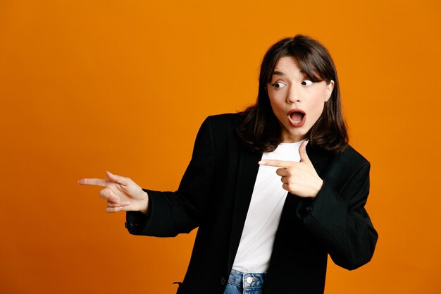 surprised points at side young beautiful female wearing black jacket isolated on orange background