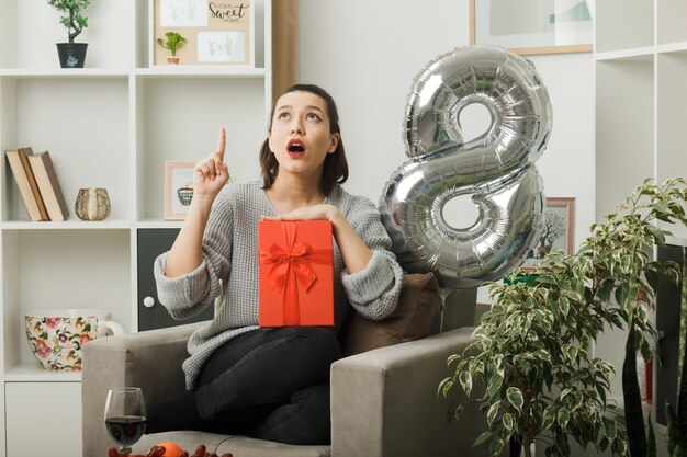 Surprised points at up beautiful woman on happy women day holding present sitting on armchair in living room