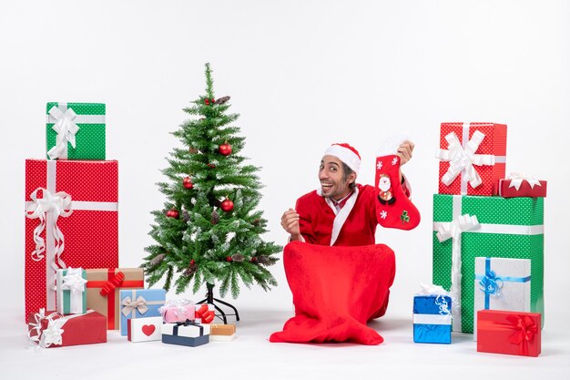 Surprised positive excited santa claus sitting on the ground and holding christmas sock near gifts and decorated new year tree on white background