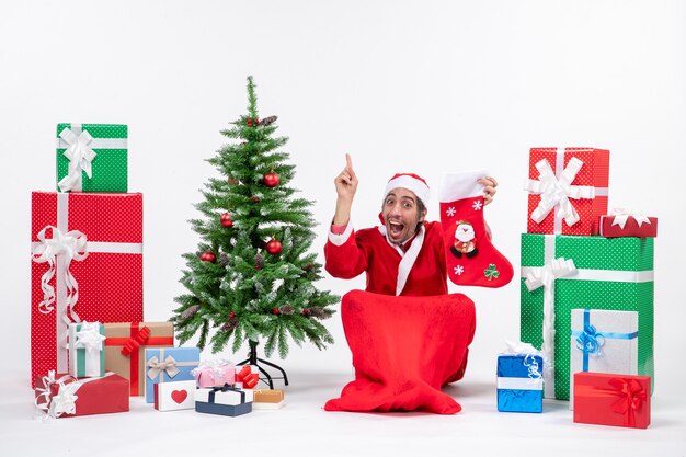 Surprised positive funny santa claus pointing above sitting on the ground and holding christmas sock near gifts and decorated new year tree on white background