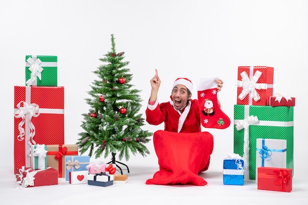 Surprised positive funny santa claus pointing above sitting on the ground and holding christmas sock near gifts and decorated new year tree on white background