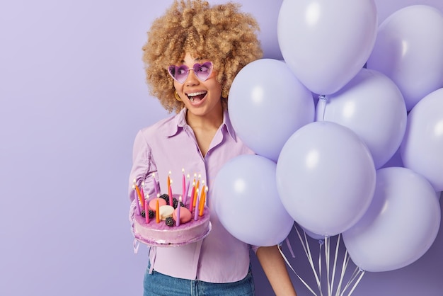 Бесплатное фото Удивленная позитивная женщина с вьющимися волосами держит кучу надутых воздушных шаров и праздничный торт празднует годовщину или особый случай, изолированный на фиолетовом фоне пустое место для рекламы