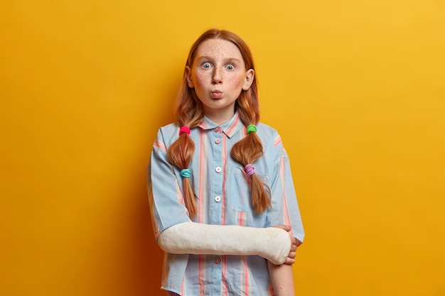 Surprised preteen girl pouts lips and looks with bugged eyes, makes funny grimace and foolishes around, wears gypsum on broken arm after accident on road. Children, face expressions