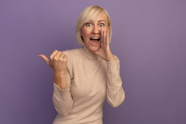 Удивленная симпатичная славянская блондинка держит руку близко ко рту, указывая на сторону, изолированную на фиолетовой стене