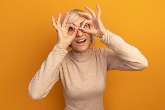 Удивленная симпатичная славянская блондинка смотрит в камеру сквозь пальцы на оранжевом