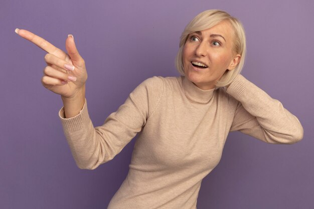 Удивленная симпатичная славянская белокурая женщина кладет руку на голову, глядя и указывая в сторону на фиолетовом