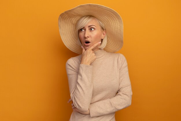 Surprised pretty blonde slavic woman with beach hat holds chin looking at side on orange