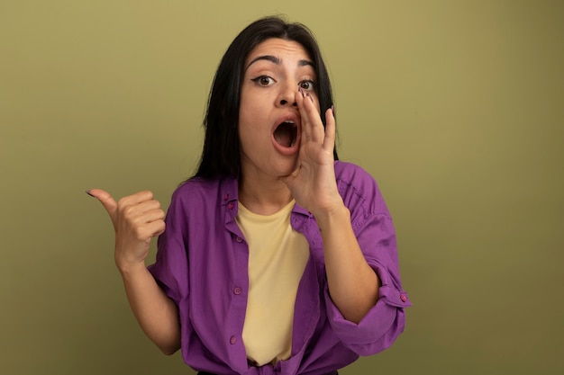 Surprised pretty brunette woman holds hand close to mouth and points at side isolated on olive green wall