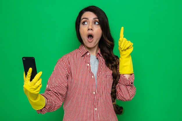 Удивленная симпатичная кавказская уборщица с резиновыми перчатками держит телефон и показывает вверх, глядя в сторону