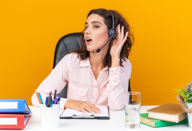 Free photo surprised pretty caucasian female call center operator on headphones sitting at desk with office tools keeping hand close to her ear trying to hear and looking at side