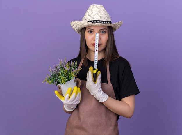 Surprised pretty caucasian female gardener wearing gardening hat holding flowerpot and tape measure