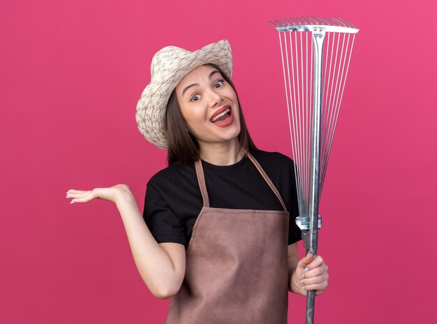 Surprised pretty caucasian female gardener wearing gardening hat holds leaf rake and keeps hand open isolated on pink wall with copy space