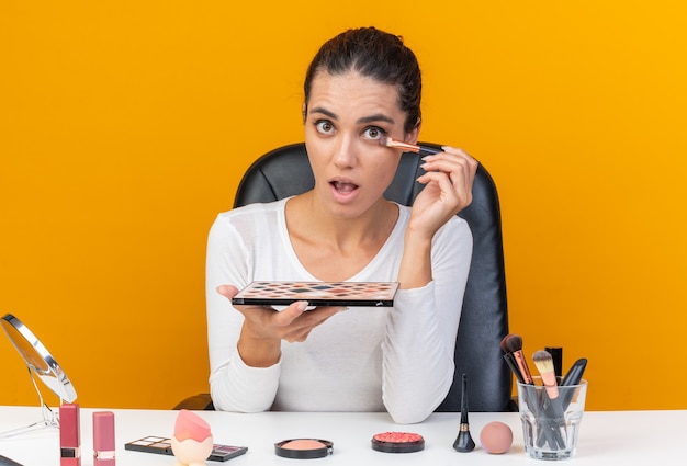 Free photo surprised pretty caucasian woman sitting at table with makeup tools holding eyeshadow palette and applying eyeshadow isolated on orange wall with copy space