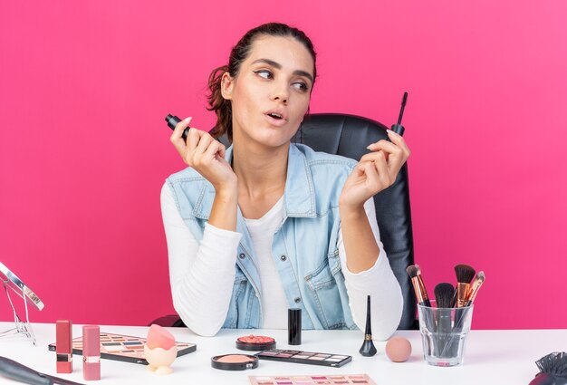 Surprised pretty caucasian woman sitting at table with makeup tools holding and looking at eyeliner isolated on pink wall with copy space