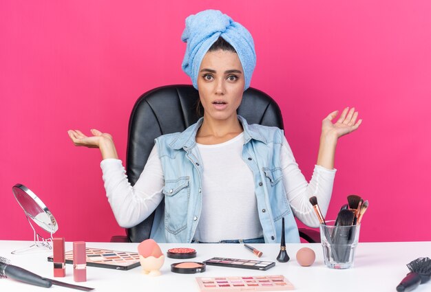 Surprised pretty caucasian woman with wrapped hair in towel sitting at table with makeup tools keeping hands open