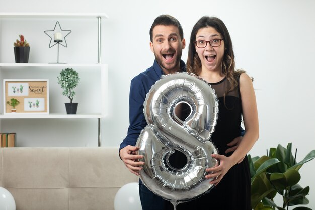 Surprised pretty couple holding balloon shaped eight standing in living room on march international women's day