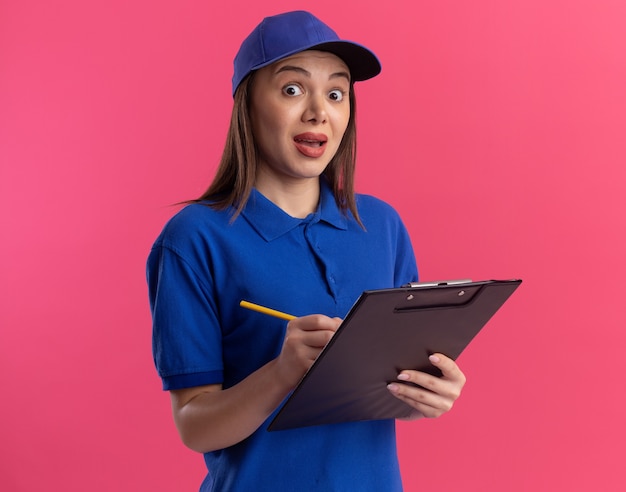 Surprised pretty delivery woman in uniform holds pencil and clipboard on pink