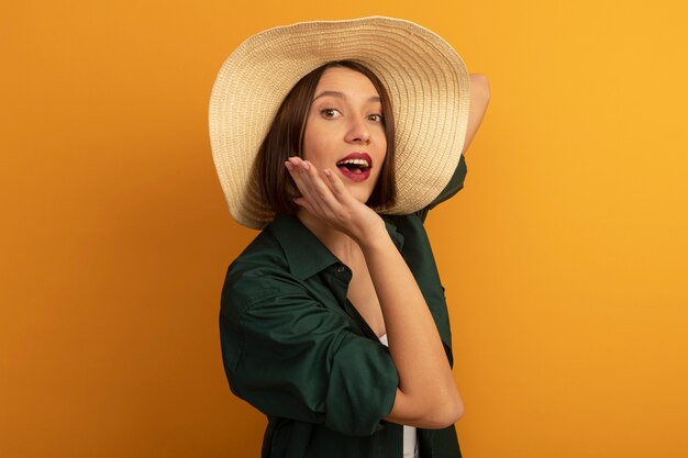 Surprised pretty woman with beach hat puts hand on chin and looks at front isolated on orange wall