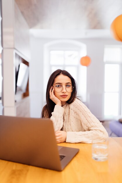 Surprised pretty woman with lond black hair is working on her laptop and sitting at home at the wooden table