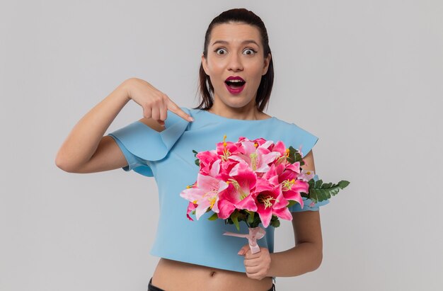 Surprised pretty young woman holding and pointing at bouquet of flowers