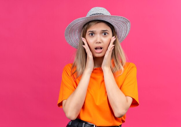 A surprised pretty young woman in an orange t-shirt wearing sunhat keeping hands on face while looking on a pink wall