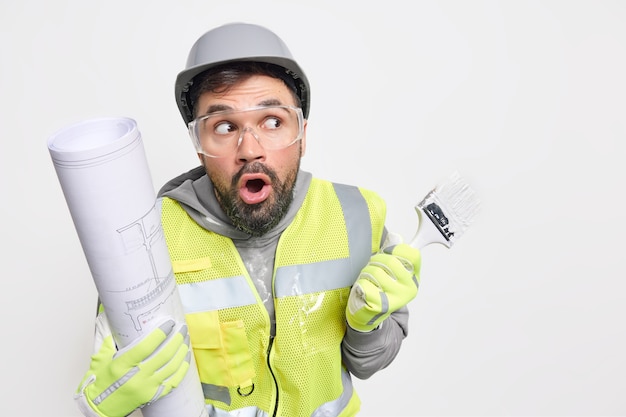 Surprised professional industry engineer poses with architectural project holds paint brush redecorates house accodting to design plan stands 