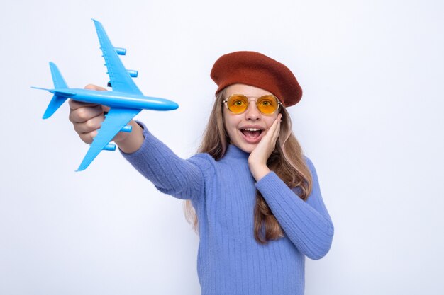 Surprised putting hand on cheek beautiful little girl wearing glasses with hat holding out toy airplane