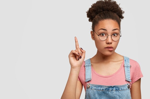 Surprised puzzled African American lady bites lower lip, points with index fingers upwards, suggests looking up, looks with uncertainty, wears optical glasses, stands against white wall, copy space