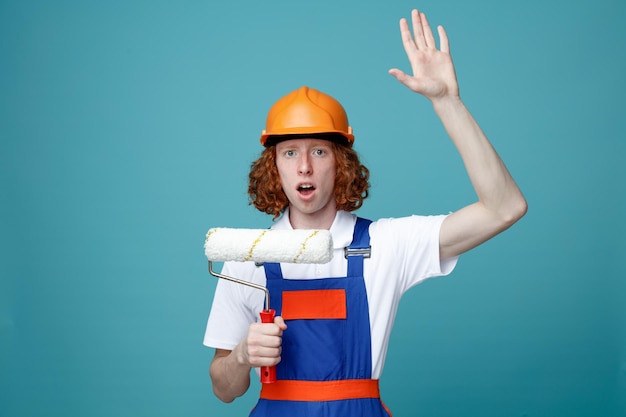 Surprised raising hand young builder man in uniform holding roller brush isolated on blue background