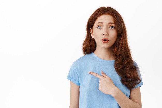 Surprised redhead girl asking question and pointing left, showing something curious aside, looking at camera questioned and intrigued, standing over white background