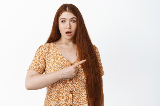 Surprised redhead girl asks question and points right at product advertisement showing banner standing curious against white background