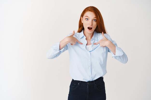 Surprised redhead girl student pointing at herself, open mouth and stare at front wondered, white wall