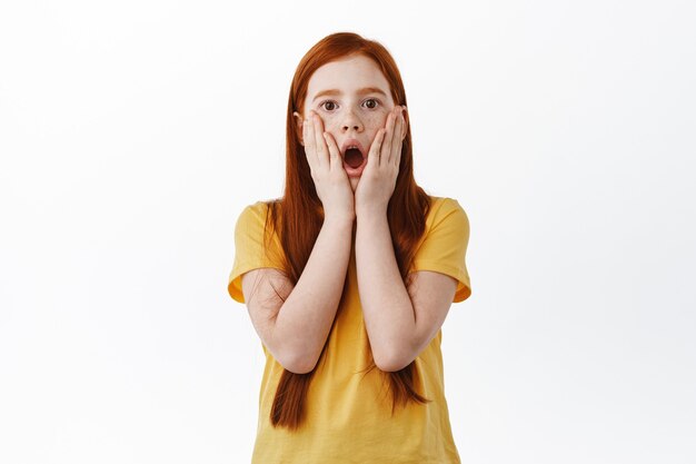 Surprised redhead girl with freckles gasping, drop jaw and stare impressed at front, holding hands on face startled and amazed, white wall