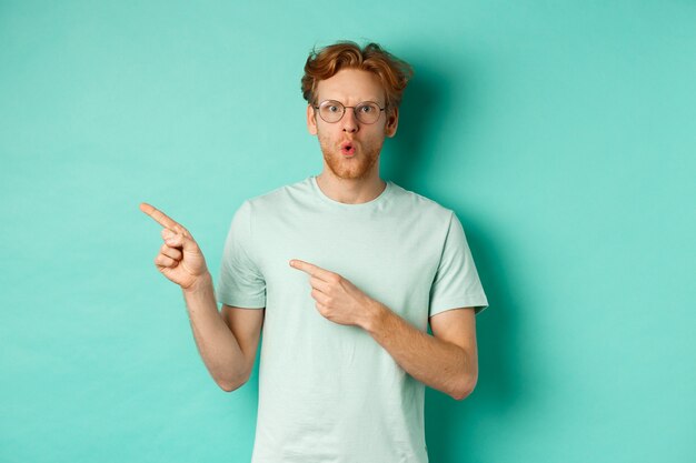 Surprised redhead guy in glasses and t-shirt pointing fingers left, saying wow and showing promo offer, checking out special deal, standing over turquoise background.