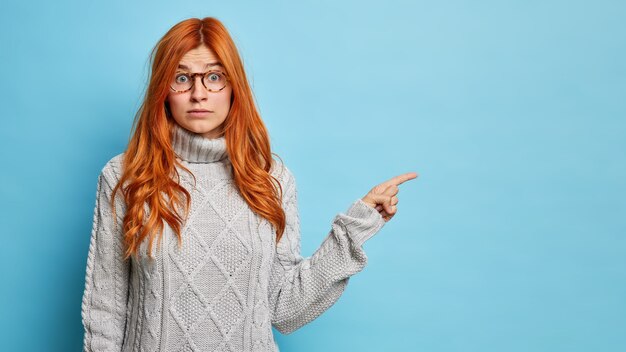 Surprised redhead woman sees incredible thing points away on copy space dressed in knitted sweater.