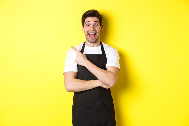 Free photo surprised salesman in black apron pointing finger upper left corner, showing shop promo offer, standing against yellow background.