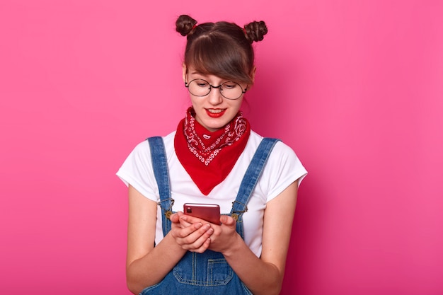 Surprised satisfied cute female typing messages on her phone, watching videos, wearing denim stylish overall