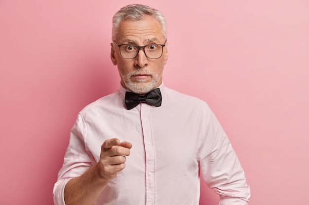 Surprised senior man wears elegant shirt with black bowtie, transparent glasses, points into camera, poses against pink background.