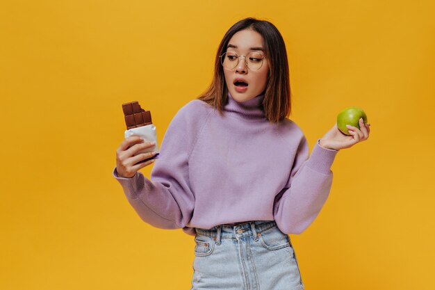 眼鏡をかけた驚いたシャツの髪の少女がミルクチョコレートバーを見る