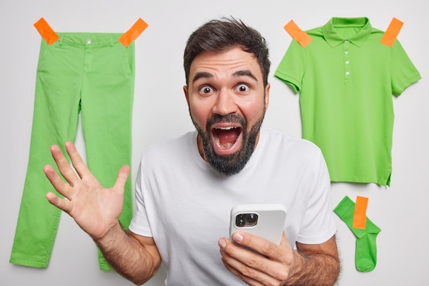 Удивленный шокированный бородатый молодой человек держит мобильный телефон, реагирует на удивительные новости, громко восклицает, одетый в повседневную простую футболку, позирует в помещении с зеленой одеждой, висящей сзади, продает одежду онлайн