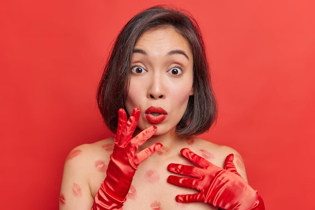 Удивленная шокированная брюнетка азиатская женщина потрясенно смотрит в камеру, носит минимальный макияж, задыхается от чудо-стендов с обнаженными плечами, имеет следы помады на теле, изолированные на ярко-красном фоне