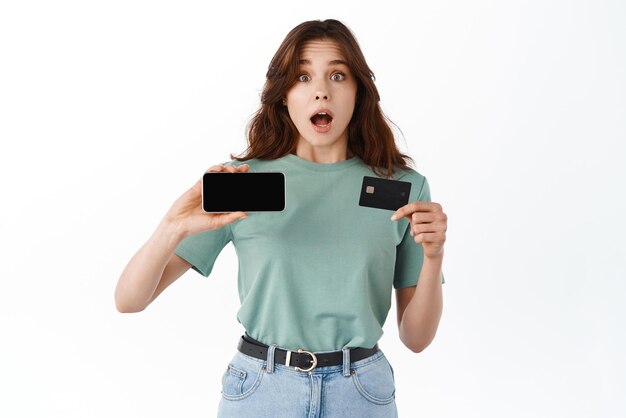 Удивленная или шокированная брюнетка, задыхаясь, показывает горизонтальный экран смартфона и пластиковую кредитную карту, демонстрируя банковский счет, стоящий на белом фоне